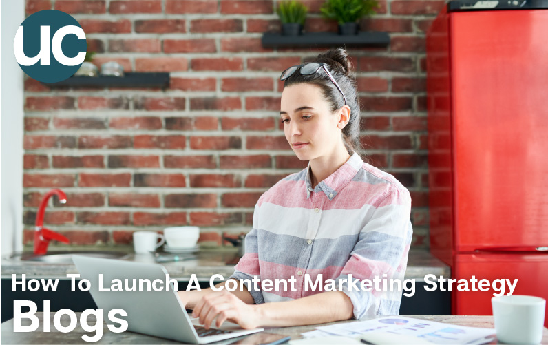How To Launch A Content Marketing Strategy - Blogs: Featured Image - A woman sits in front of her laptop to begin typing her blog. There is a red brick wall behind her.