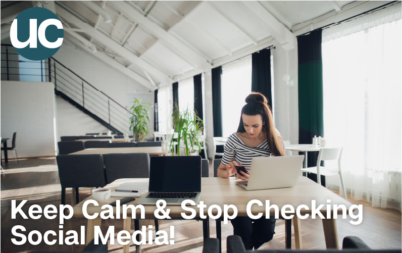 Keep Calm & Stop Checking Social Media - Featured Image: A woman sits in a large atrium coworking space at her desk and absentmindedly checks her phone during work.